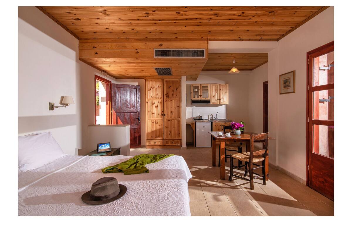Een slaapkamer met een bed, nachtkastje, lamp en hoed. Er is een houten plafond en een raam met uitzicht.