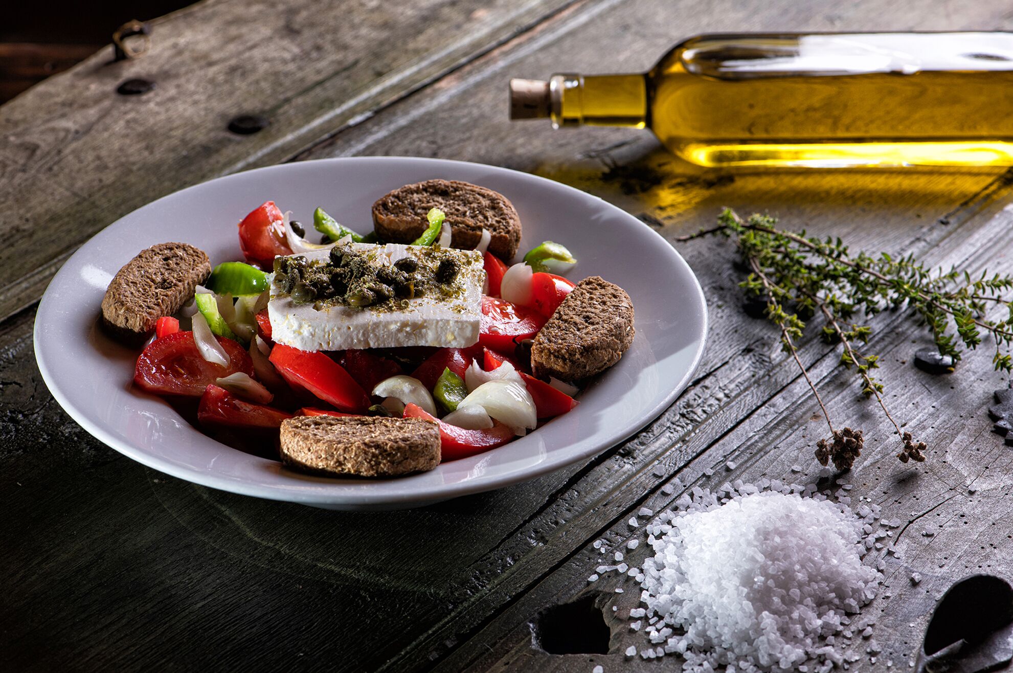 Un plato de comida con una fuente blanca sobre una mesa de madera, una botella de aceite y algunas hierbas.