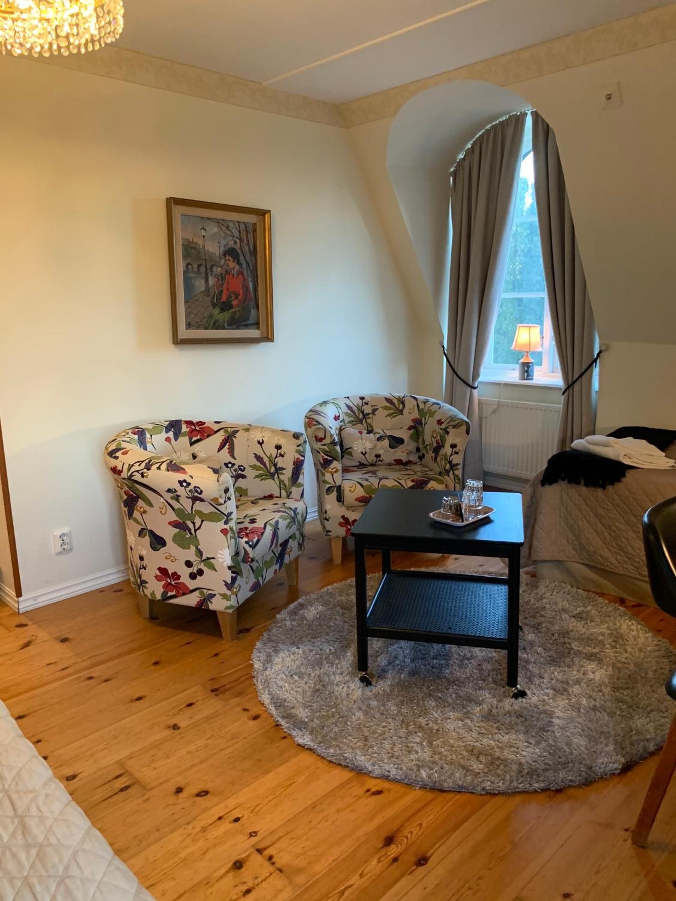 Ein gemütliches Zimmer mit einem blumigen Sofa, einem kleinen Tisch mit Tablett und einem Fenster mit Vorhängen.