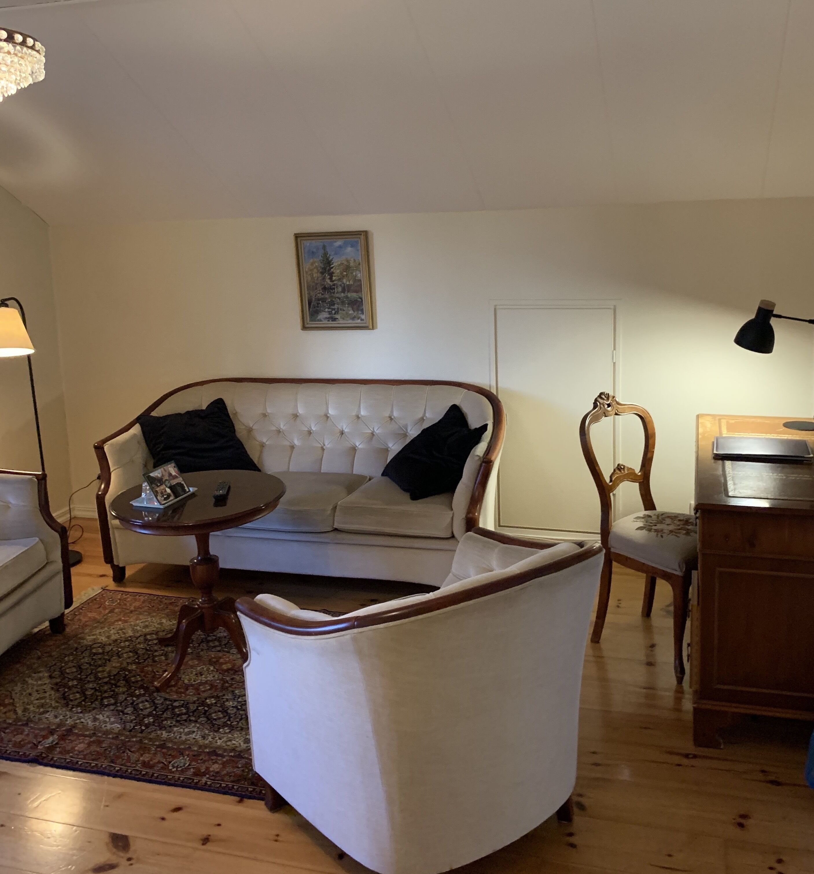 Una sala de estar con un sofá blanco, una mesa de centro redonda, un sillón de madera y un cuadro en la pared.