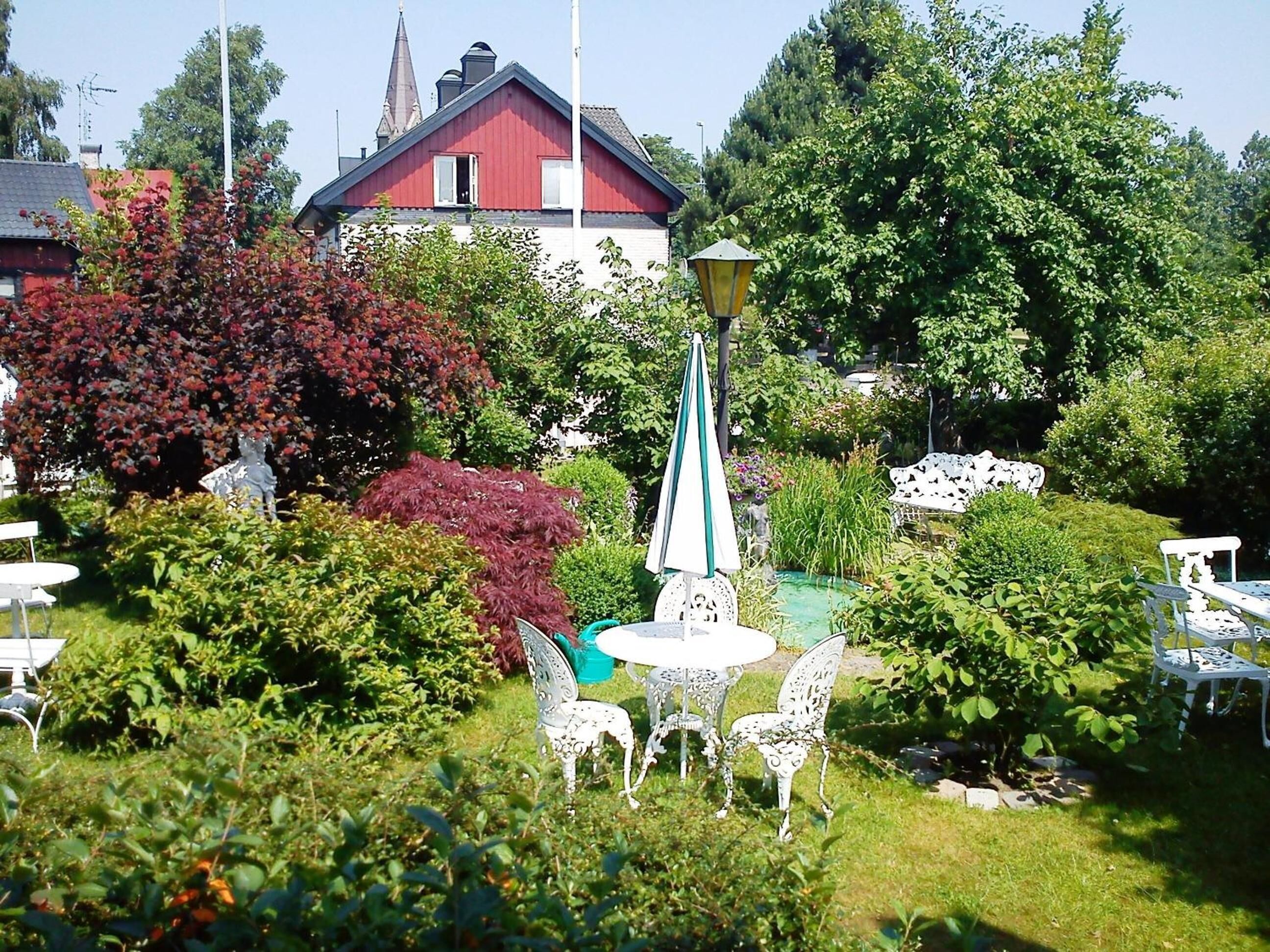 En trädgård med vita metallstolar och ett bord, omgiven av olika växter och en röd byggnad i bakgrunden.