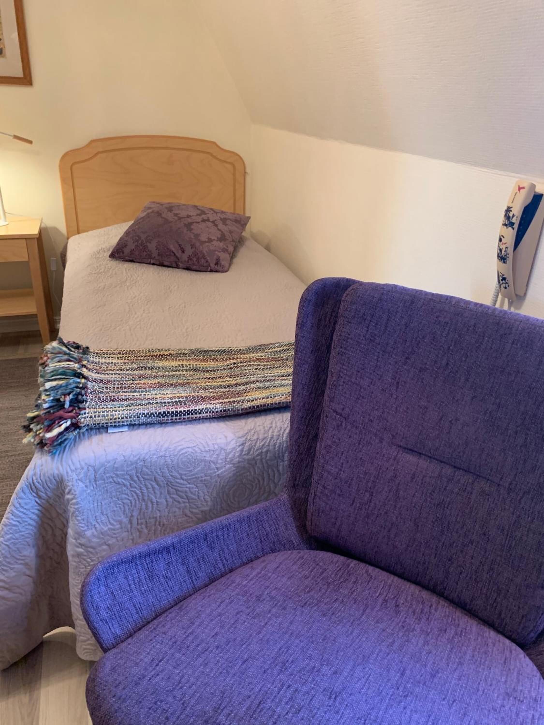 Un dormitorio con una cama, un sillón púrpura, una mesita de noche y un teléfono instalado en la pared.