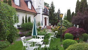 A white building with a red-tiled roof, a balcony, and a garden with white metal chairs and tables. - Nya Pallas Hotel (Falkenberg)