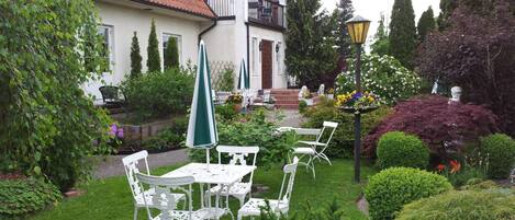 A white building with a red-tiled roof, a balcony, and a garden with white metal chairs and tables.
