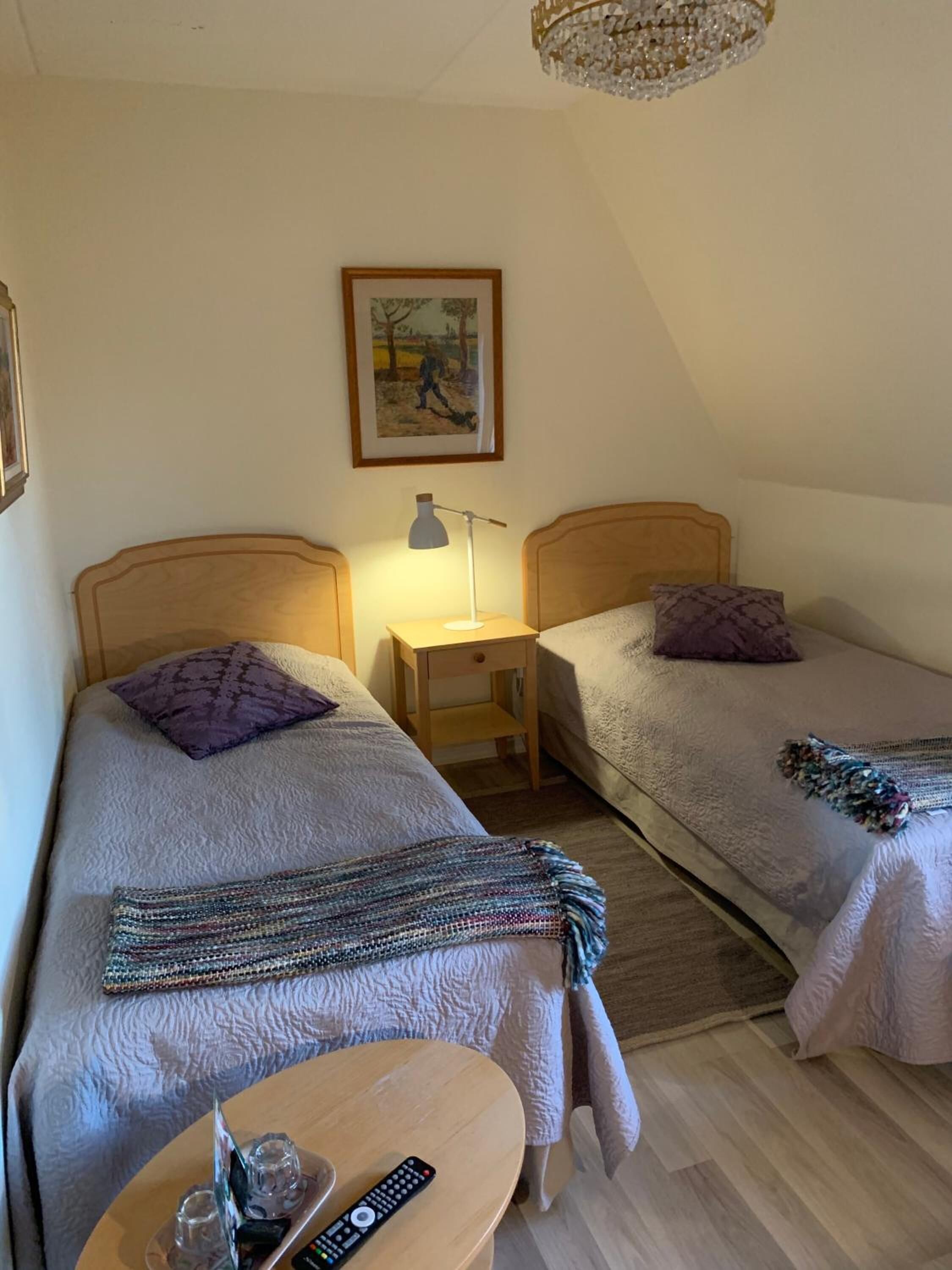 A bedroom with two single beds, a bedside table with a lamp, a small table with a remote control, and a painting on the wall.