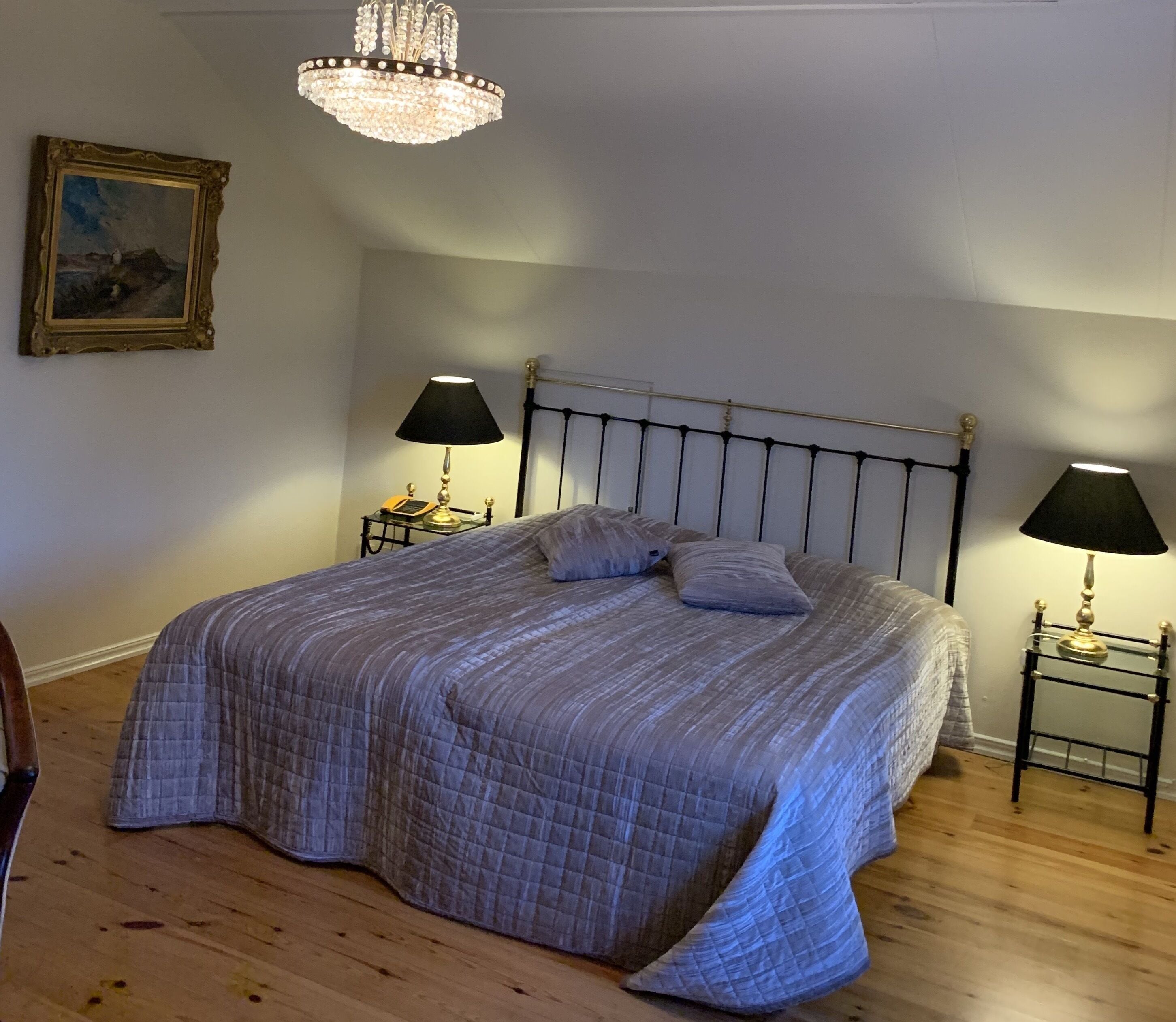 Une chambre à coucher avec un lit, deux tables de chevet avec des lampes, un lustre et un tableau encadré au mur.