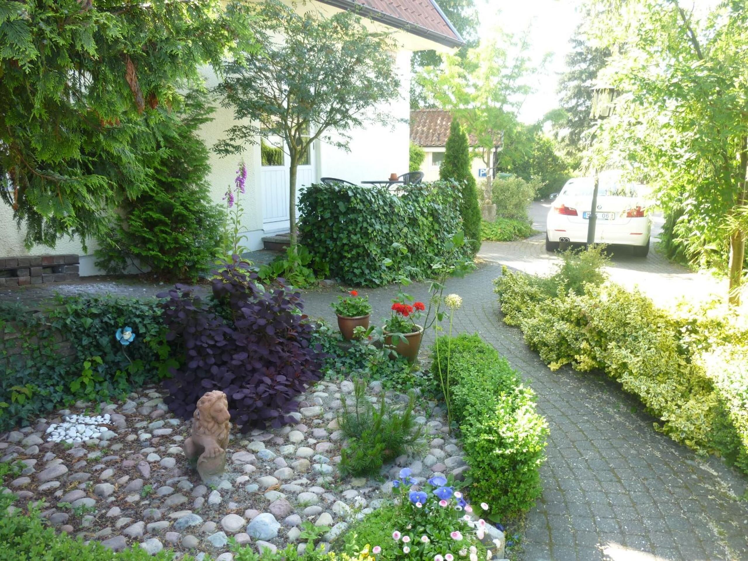 A garden with a stone statue, various plants, and a paved path leading to a white building.