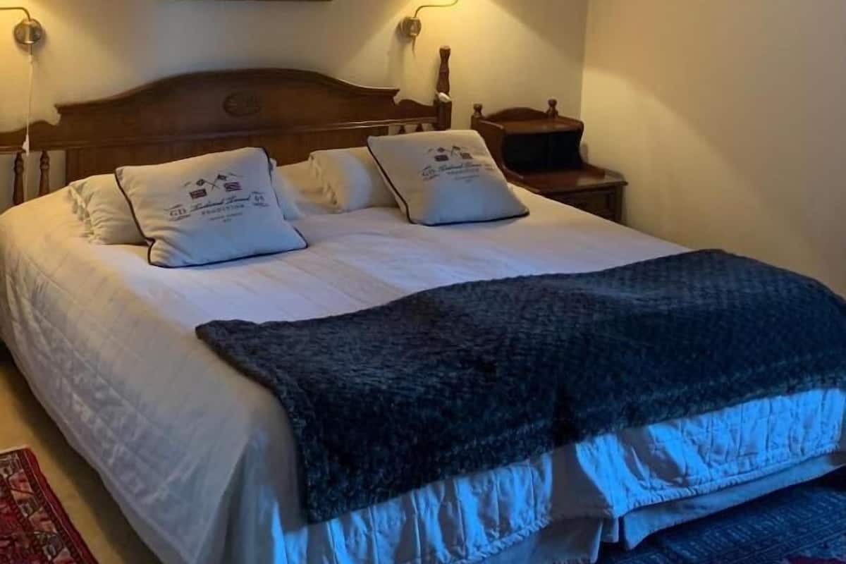 A neatly made bed with a dark blanket, two pillows, and a headboard. There is a painting on the wall above the bed and a lamp on the nightstand beside it.