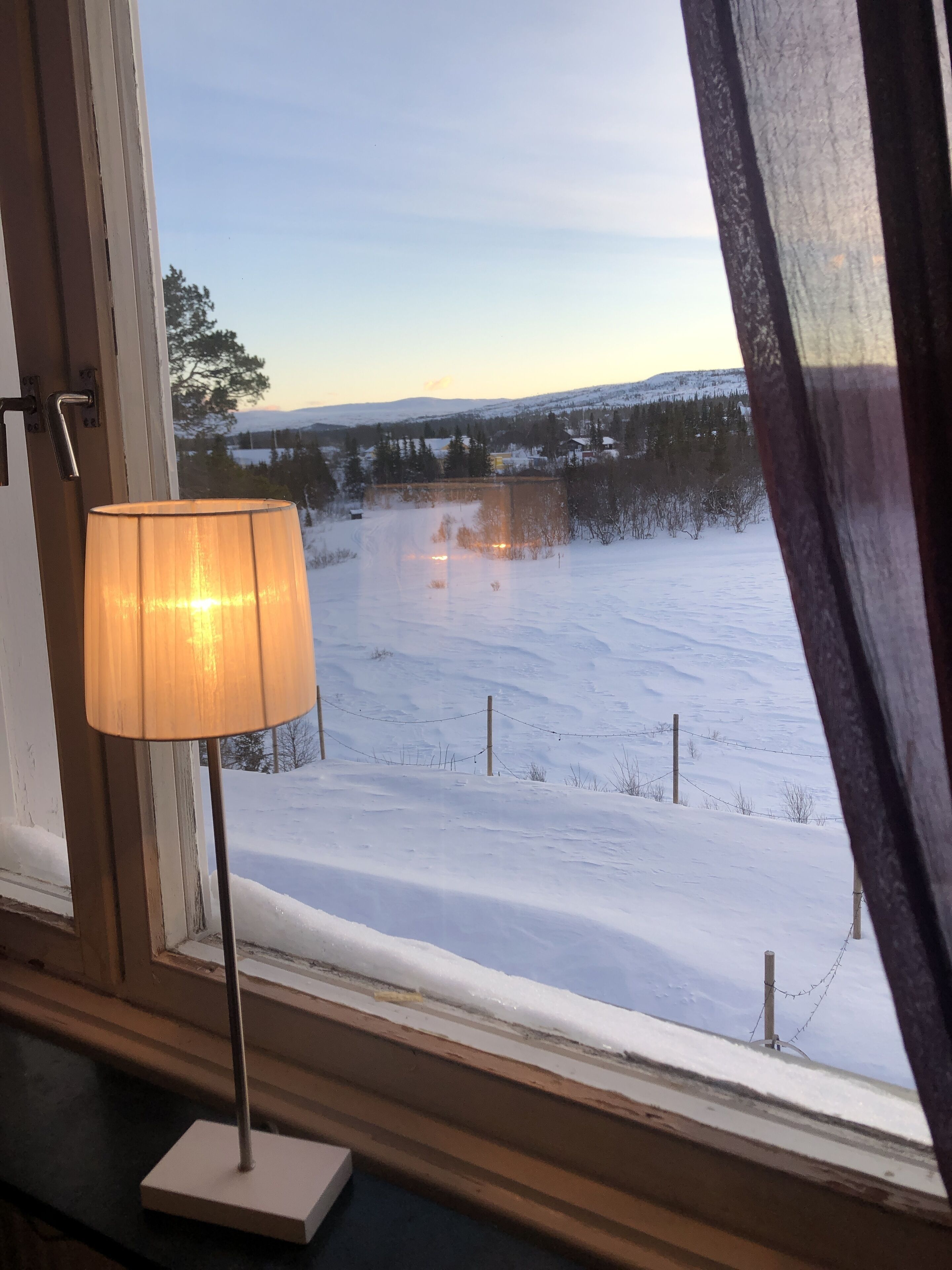 Une fenêtre donnant sur un paysage enneigé, une lampe sur une table et un rideau à moitié tiré.