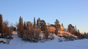 Schneebedeckte Landschaft mit einem Gebäude auf einem Hügel, umgeben von Bäumen.