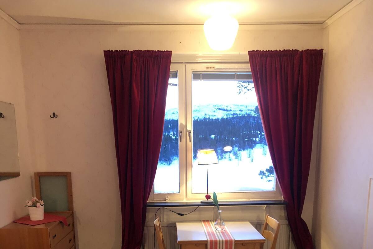 Une chambre avec deux lits, un bureau et une chaise. Il y a une fenêtre avec des rideaux, une lampe et une vue sur un paysage enneigé.