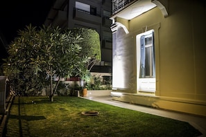 A well-lit residential building with a paved entrance, a small lawn, and a large tree. - Creta Elena (Chania)