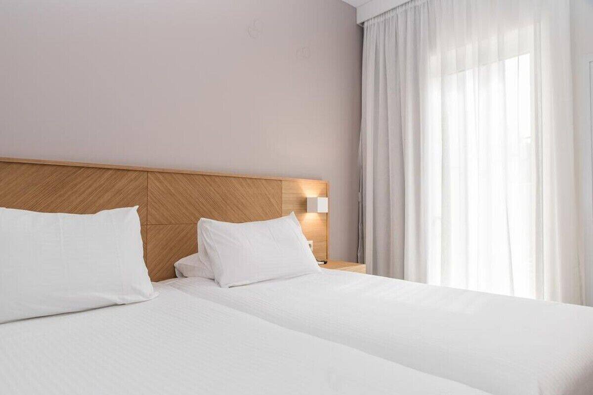 A hotel room with a wooden headboard, two white beds, a nightstand with a lamp, and a large window with sheer curtains.