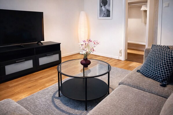 Una sala de estar moderna con una mesa de centro de vidrio, un sofá, un televisor, una lámpara y un cuadro en la pared.