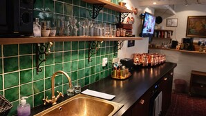 A kitchen with a copper sink, green tiled backsplash, and shelves with glassware and copper pots.
