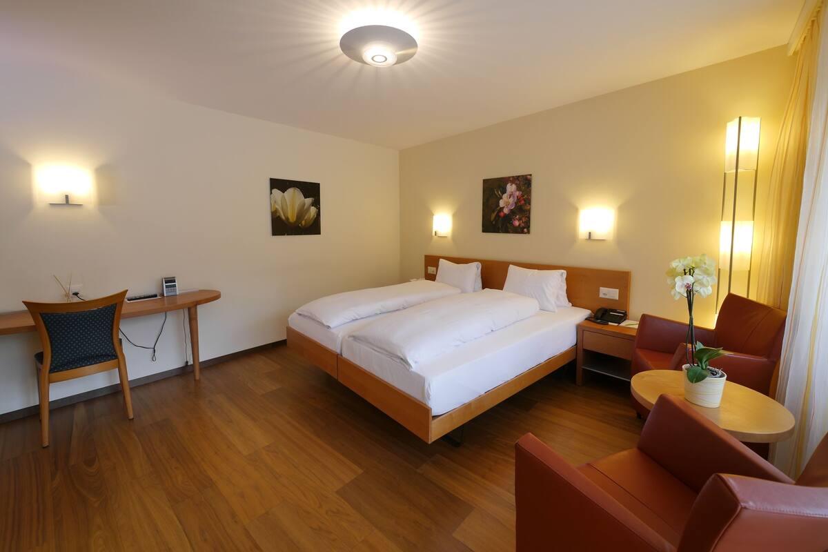 A hotel room with a large bed, a desk with a chair, a small table with a plant, and two wall-mounted lamps.