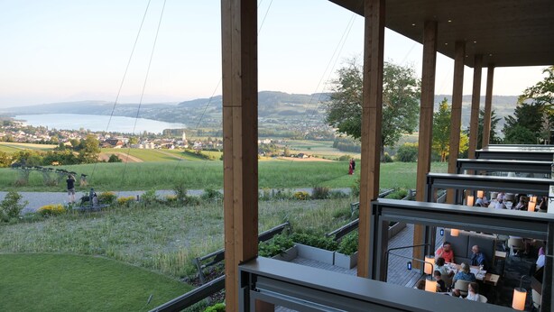 A restaurant with outdoor seating overlooking a scenic landscape, including a body of water and rolling hills.