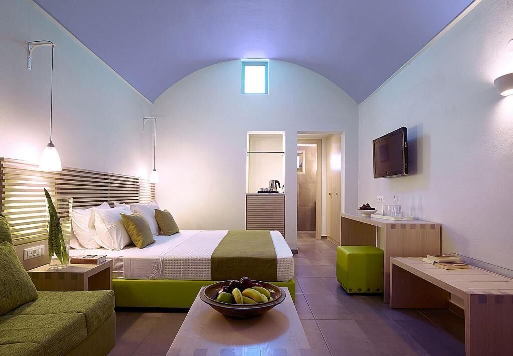 A modern hotel room with a bed, bedside table, green ottoman, and a bowl of fruit on a table.
