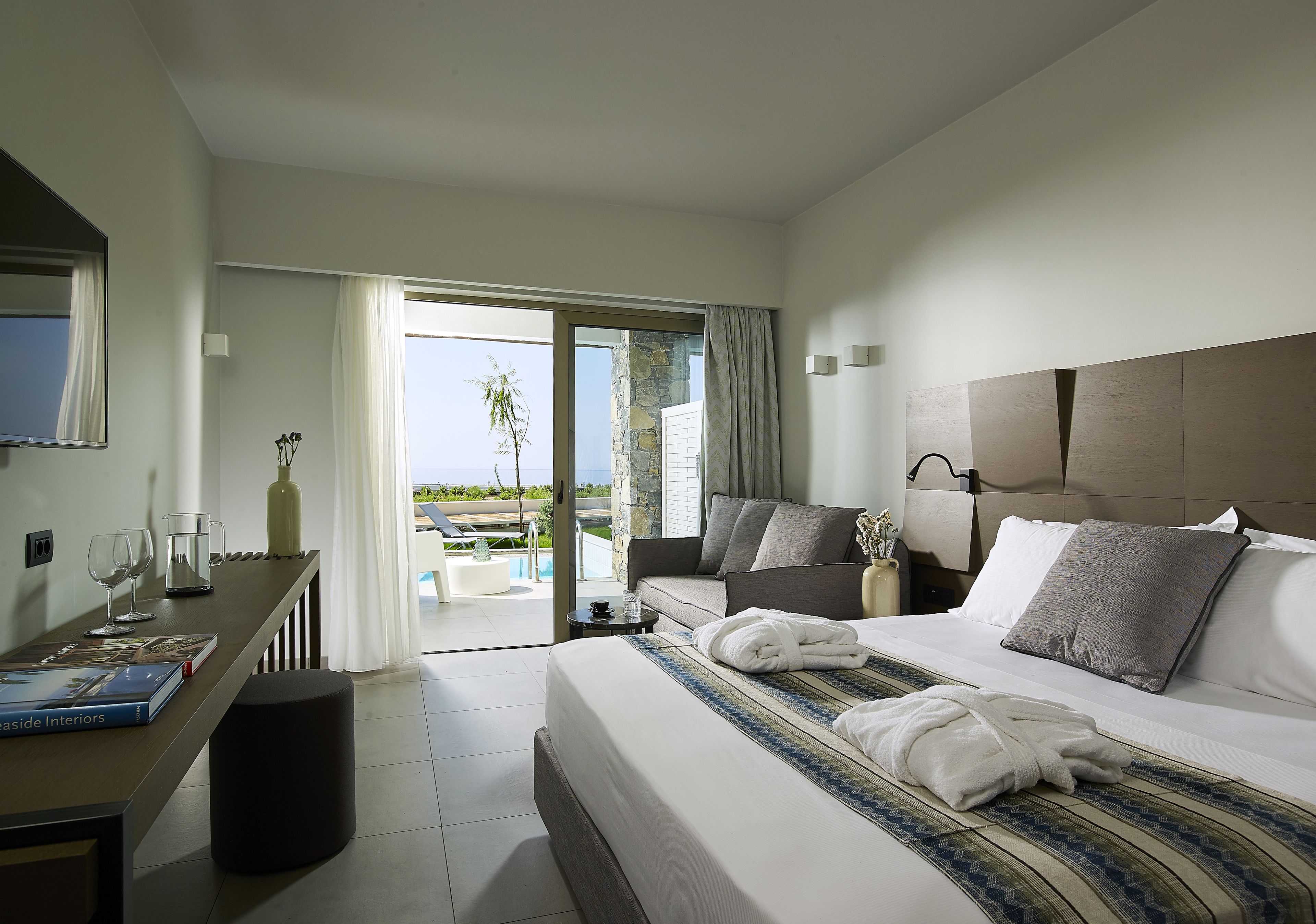 A modern hotel room with a large bed, a sofa, a dining table, and a view of the pool and greenery.