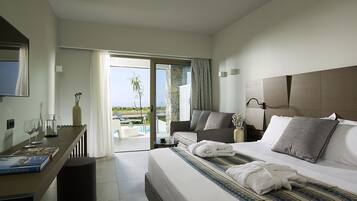 A modern hotel room with a large bed, a sofa, a dining table, and a view of the pool and greenery.