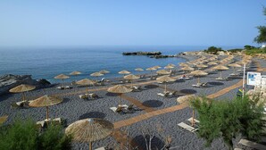 Ein Strand mit Liegen, strohgedeckten Sonnenschirmen, einer felsigen Küstenlinie und blauem Himmel.