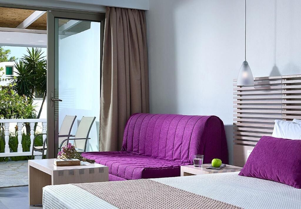 A modern hotel room with a bed, a sofa, a small table with a glass of water and an apple, and a balcony with chairs and plants.