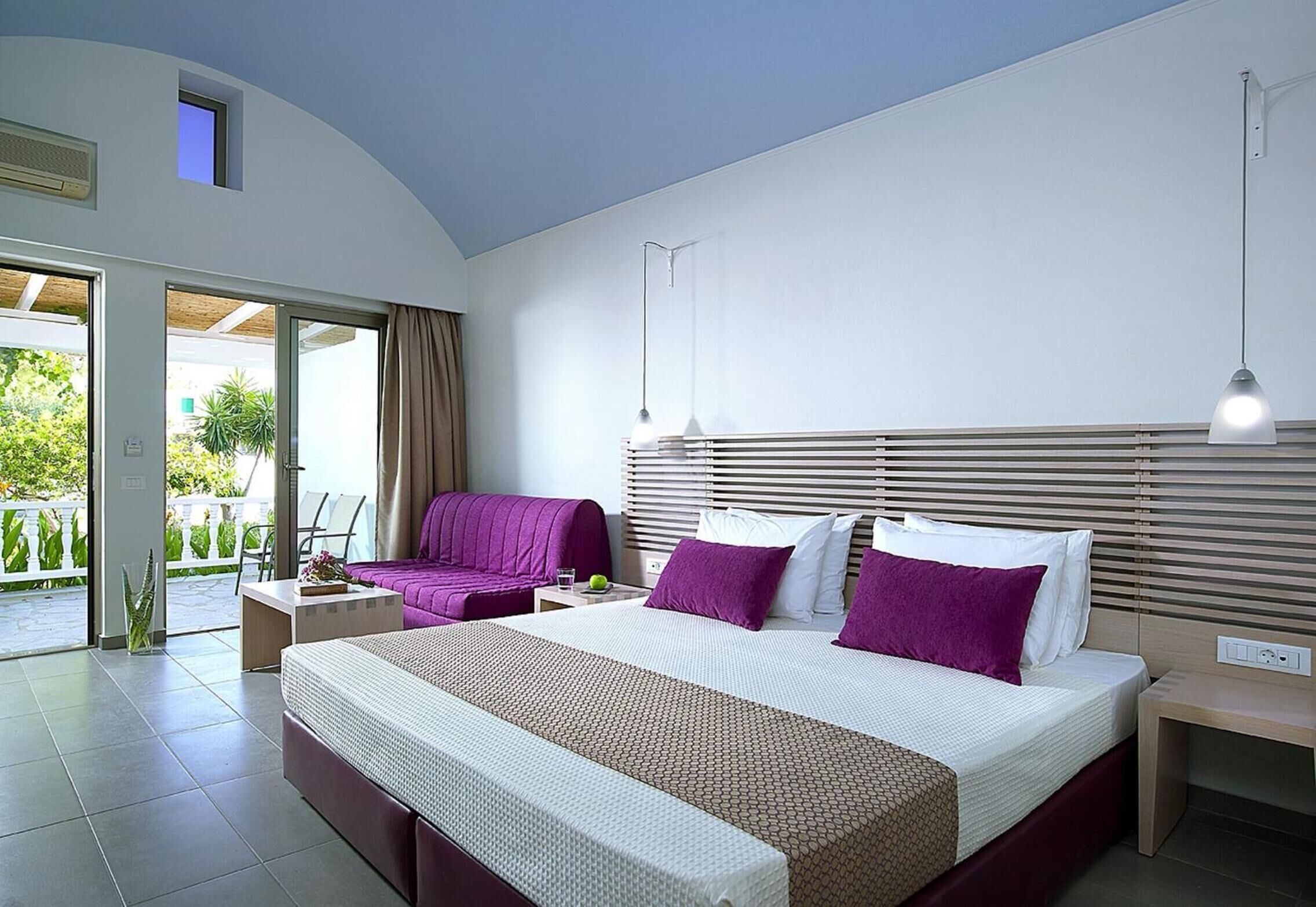 A modern hotel room with a large bed, a sofa, a desk, and a balcony with a view of greenery.