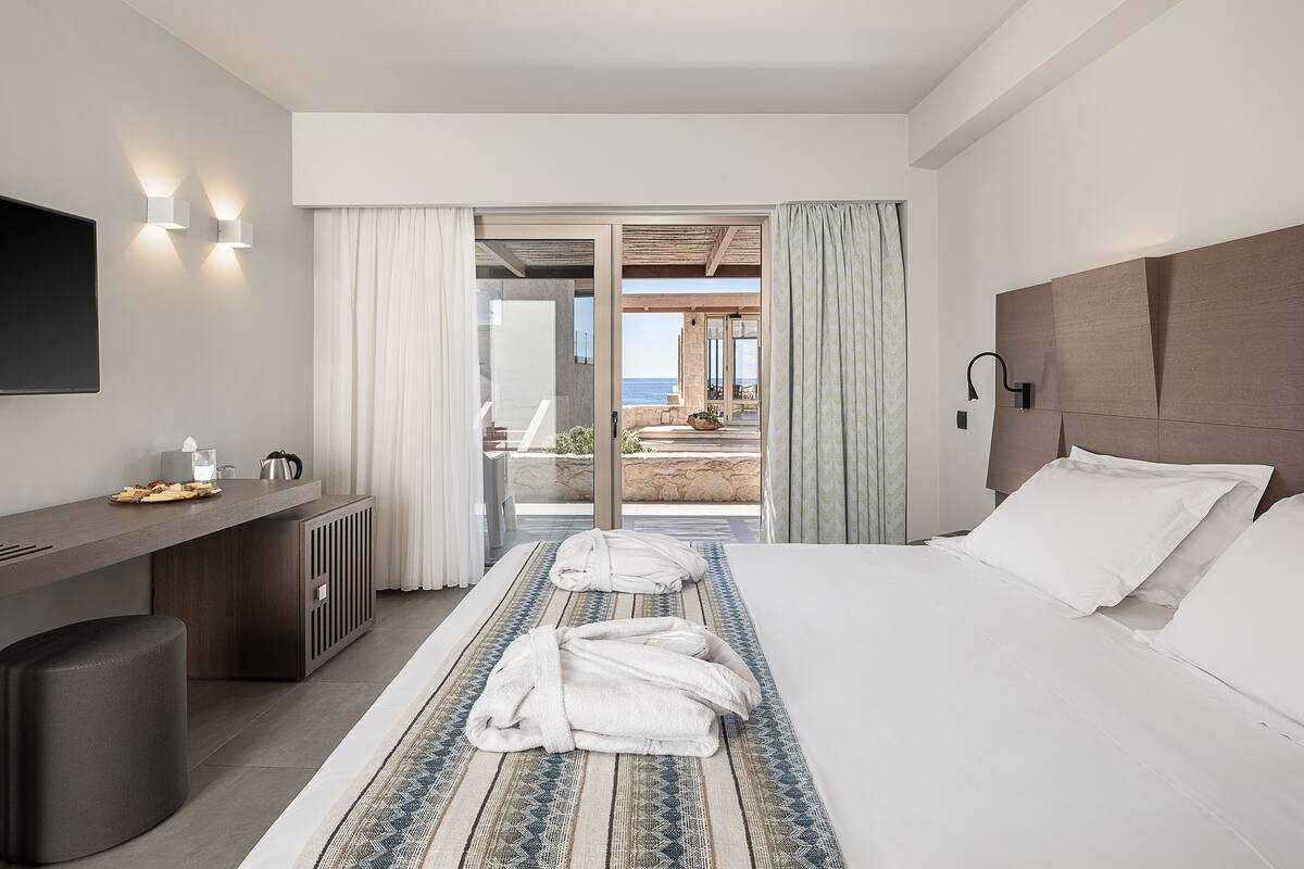 Une chambre d’hôtel moderne avec un grand lit, un bureau doté d’un téléviseur à écran plat et une vue sur la mer, visible à travers la porte vitrée.