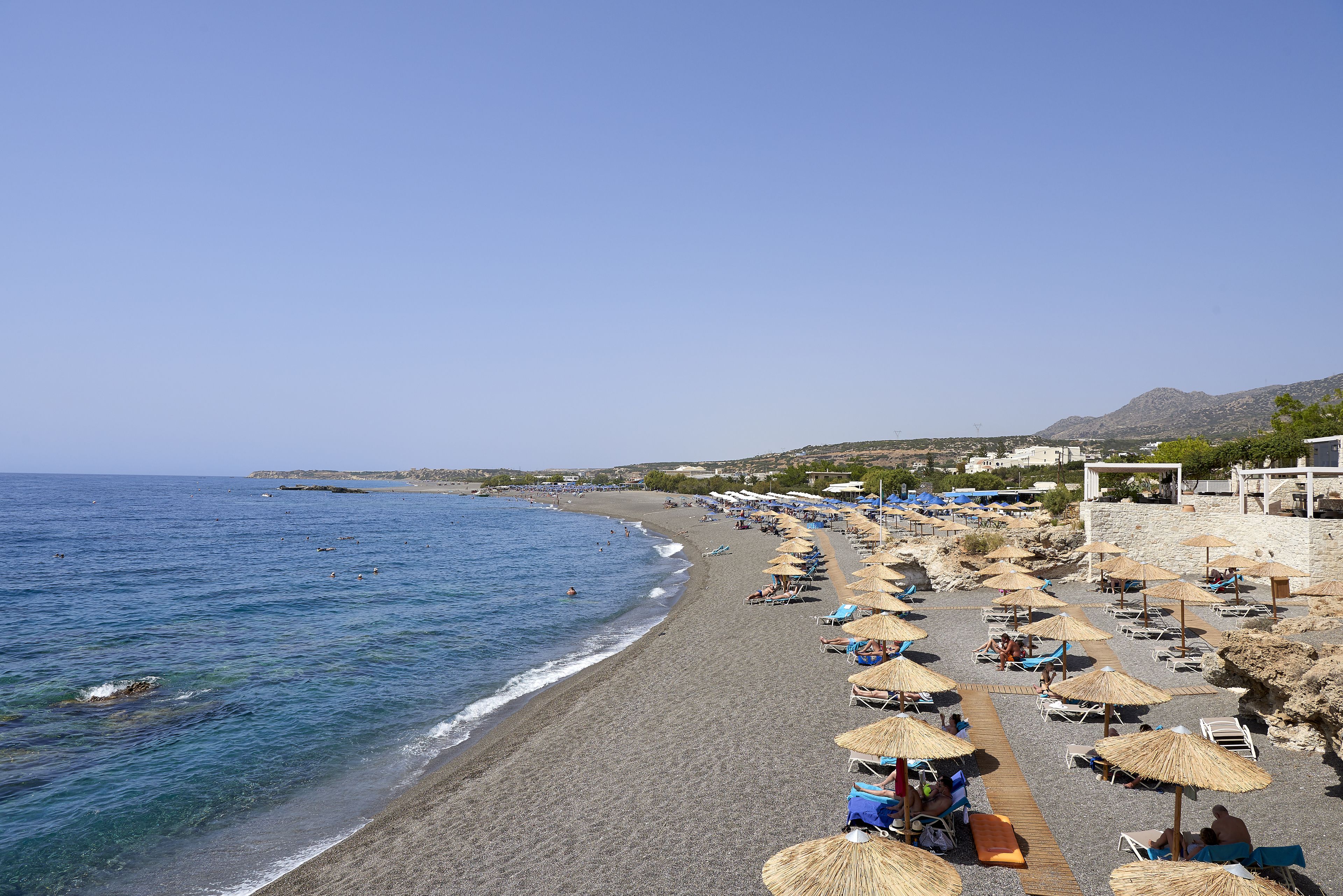 A beach with rows of sun loungers and umbrellas, a clear blue sky, and a calm sea.