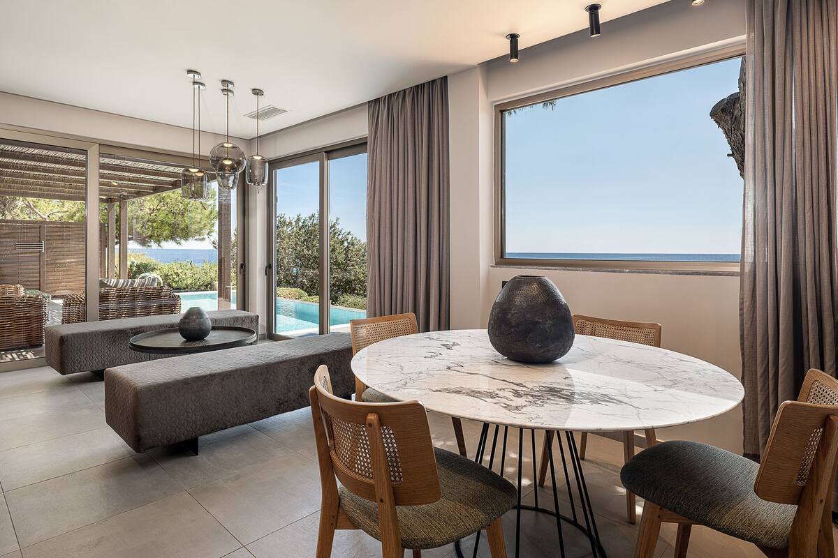 Un espace repas moderne avec une table ronde en marbre, des chaises en bois et une grande fenêtre donnant sur la mer.