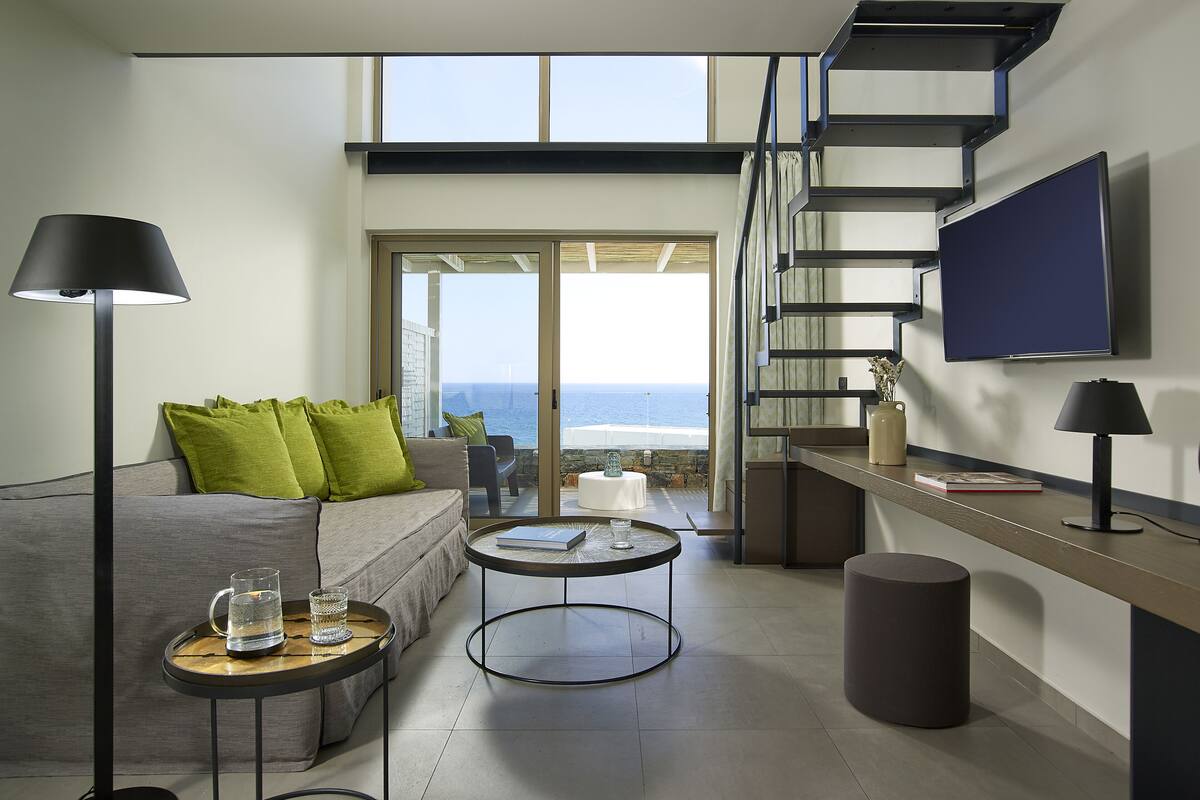 Un salon moderne comprenant un canapé, deux tables d’appoint, une table basse, un téléviseur et un escalier menant à un balcon extérieur avec vue sur la mer.