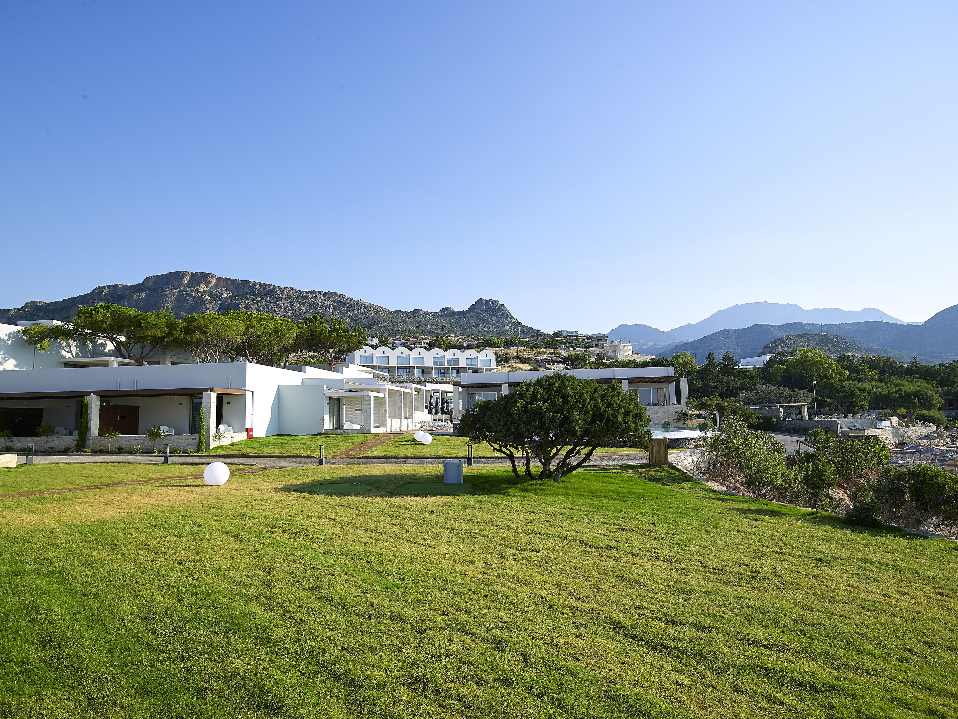 A modern hotel with multiple white buildings, a grassy lawn, and mountains in the background.