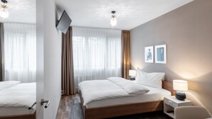 A hotel room with a wooden bed, a bedside table with a lamp, a TV mounted on the wall, and a window with sheer curtains. - Hotel Birsighof (Basel)