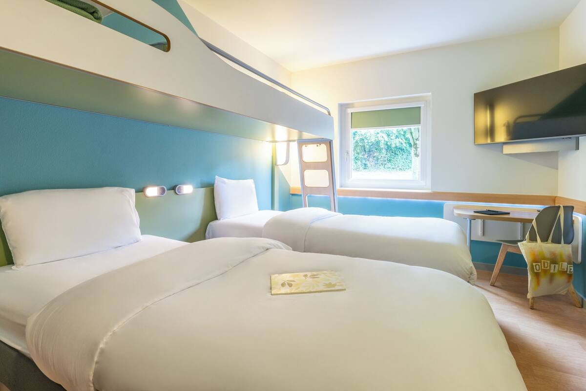 A hotel room with two beds, a desk, a chair, a TV, and a window with a view of trees.