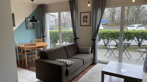 A modern living room with a sofa, a coffee table, and a dining area with a table and chairs. Large windows provide a view of the outside.