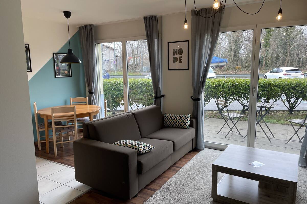 A modern living room with a sofa, a coffee table, and a dining area with a table and chairs. Large windows provide a view of the outside.