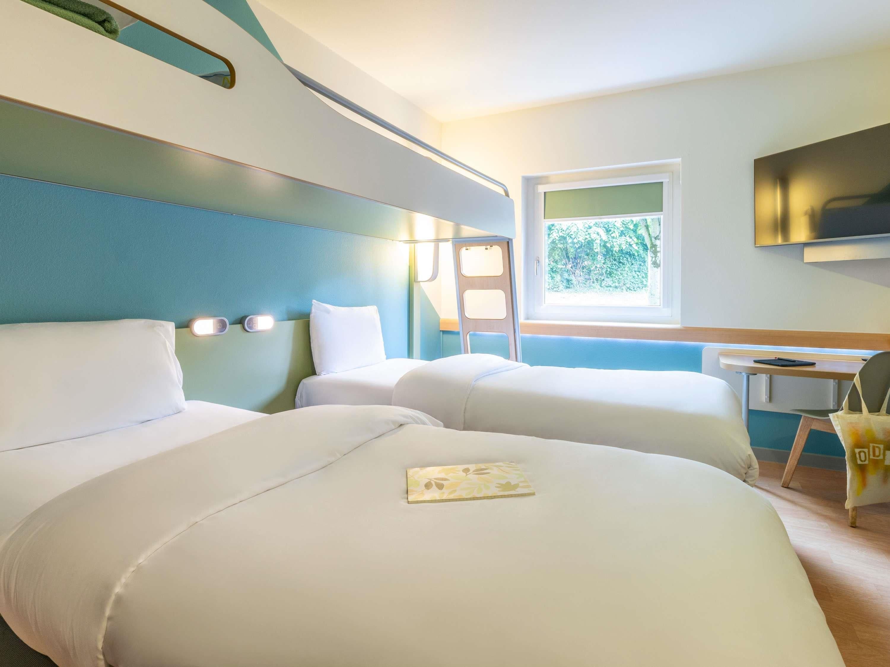 Une chambre d’hôtel avec deux lits, un bureau, une chaise et une fenêtre donnant sur les arbres.