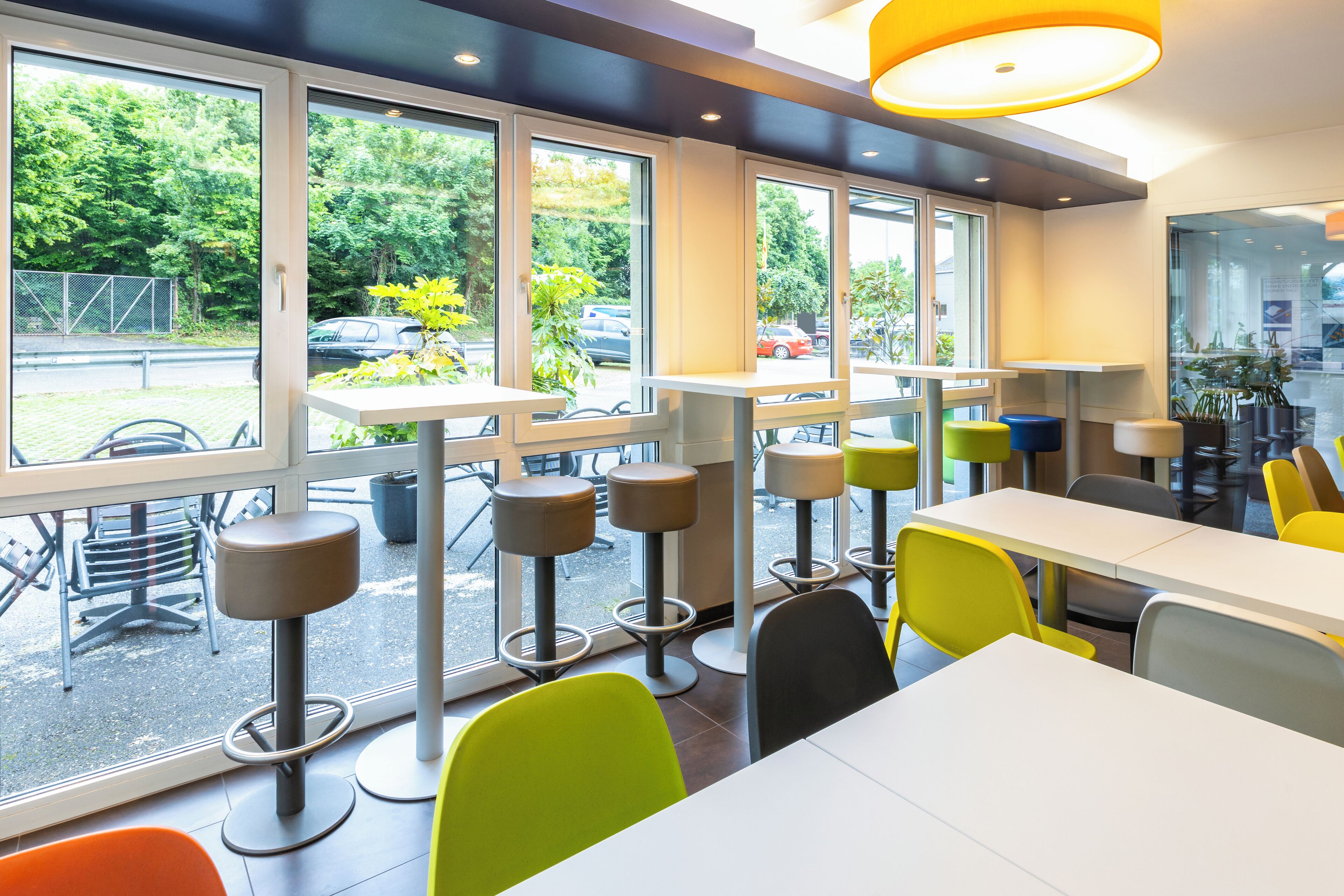 A modern indoor seating area with tables and chairs, large windows, and a view of the parking lot and greenery outside.