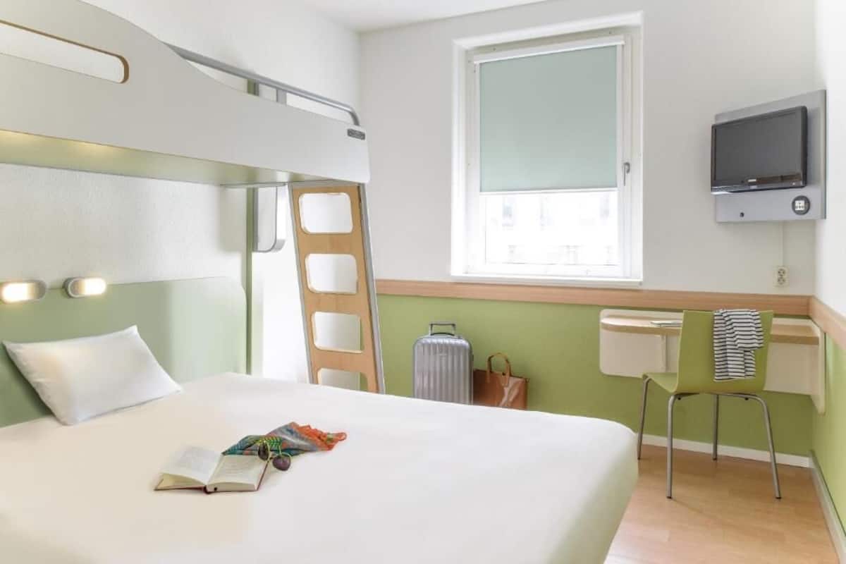 A hotel room with a bunk bed, a desk with a chair, a television, and a window with a view.