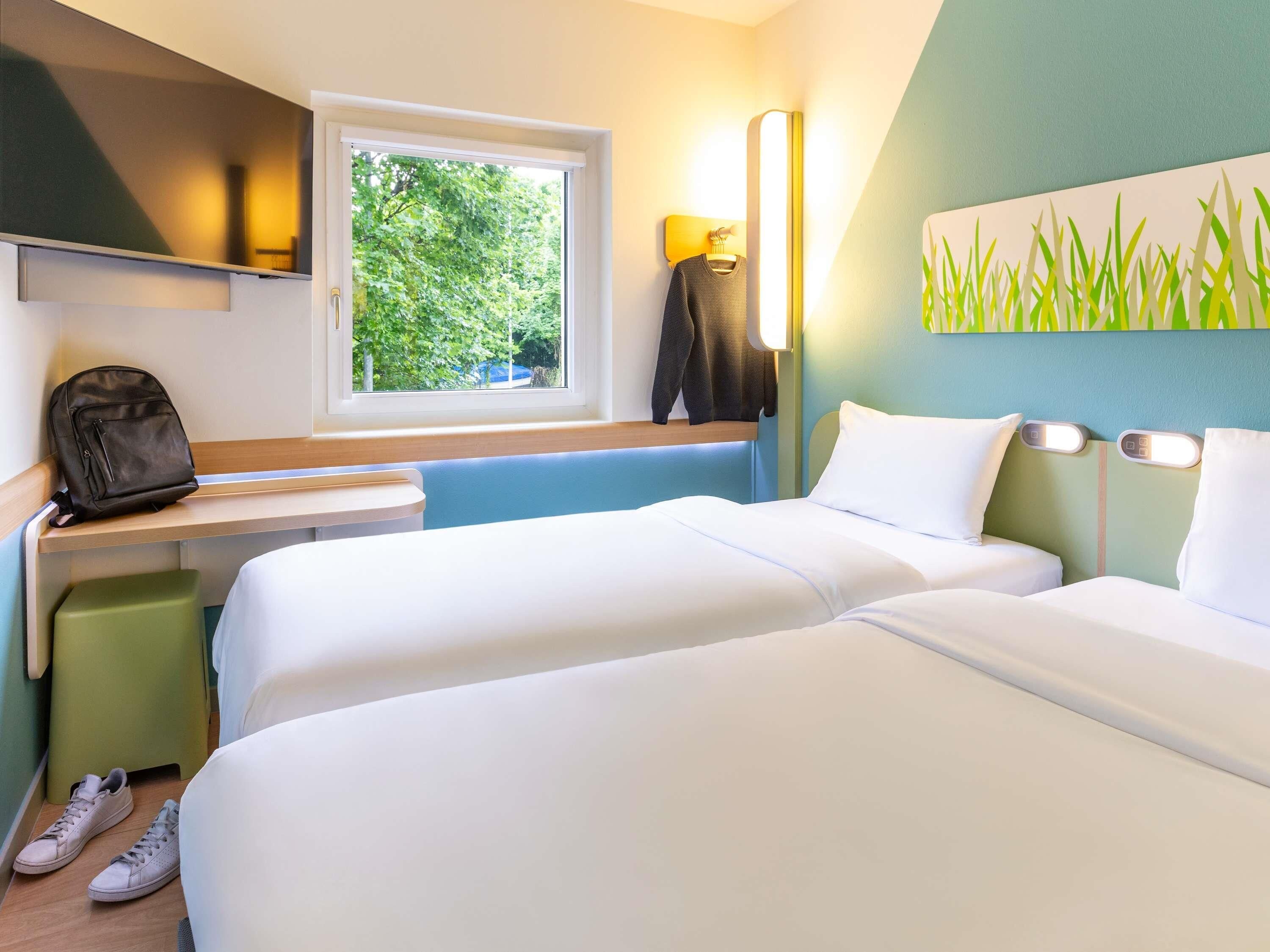 Ein Hotelzimmer mit zwei Betten, einem Fenster mit Blick auf Grünflächen, einem Schreibtisch mit einem Rucksack und einer Wandlampe.