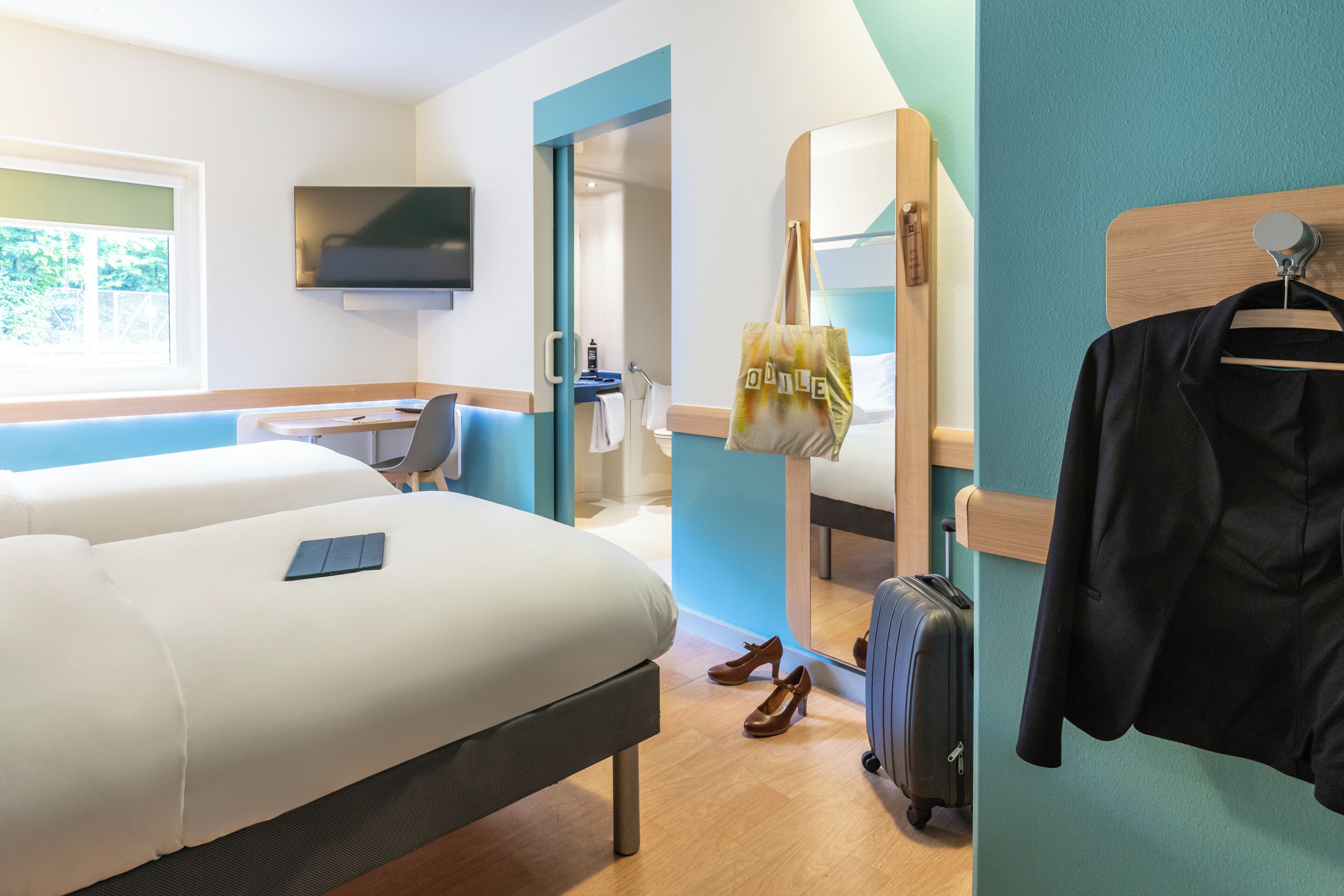 Una camera d'albergo con un letto, una scrivania, una televisione, uno specchio, una borsa appesa, un attaccapanni e una valigia.