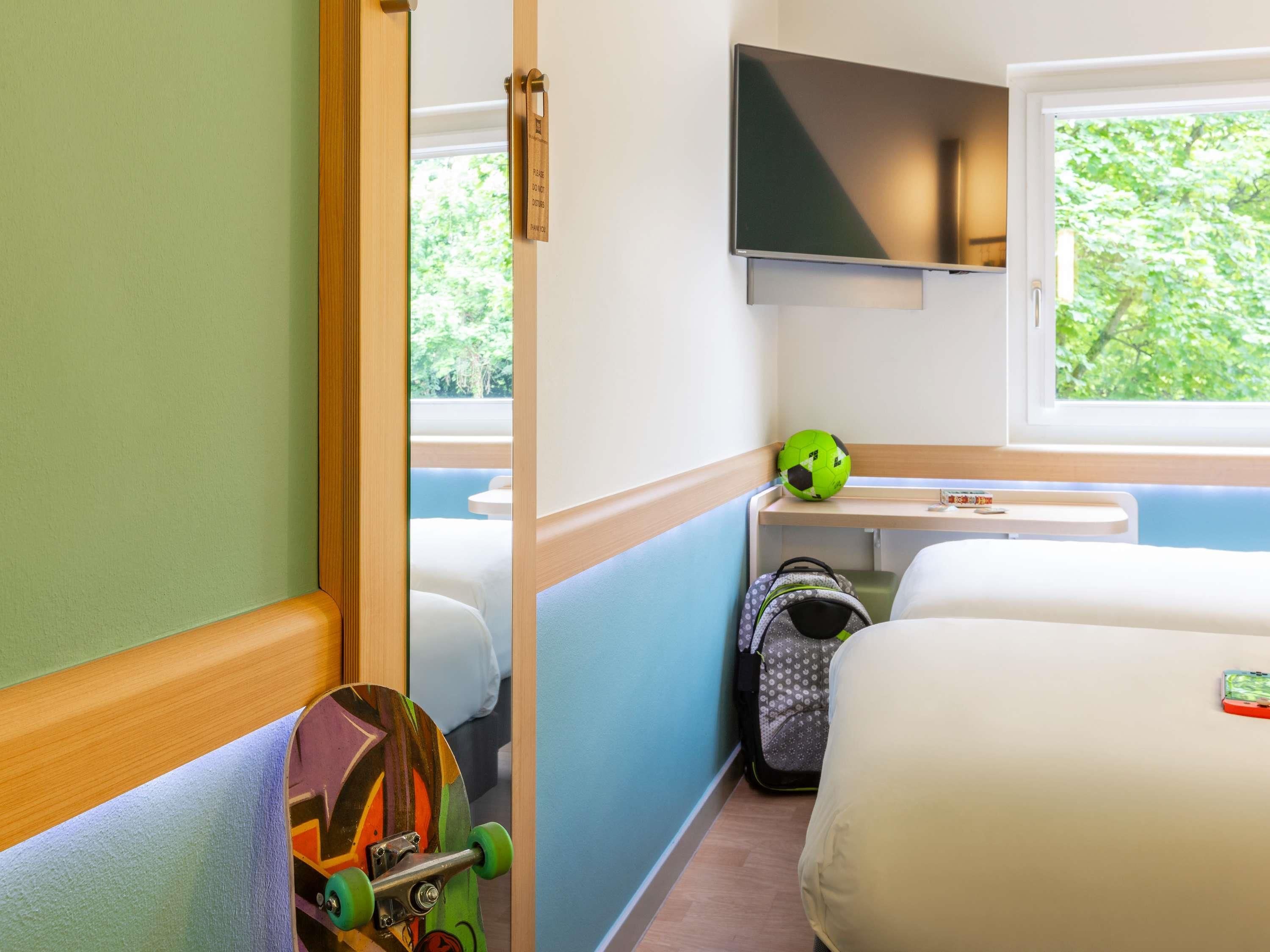 A compact room with a bed, a television mounted on the wall, a window with a view of greenery, and a skateboard leaning against the wall.