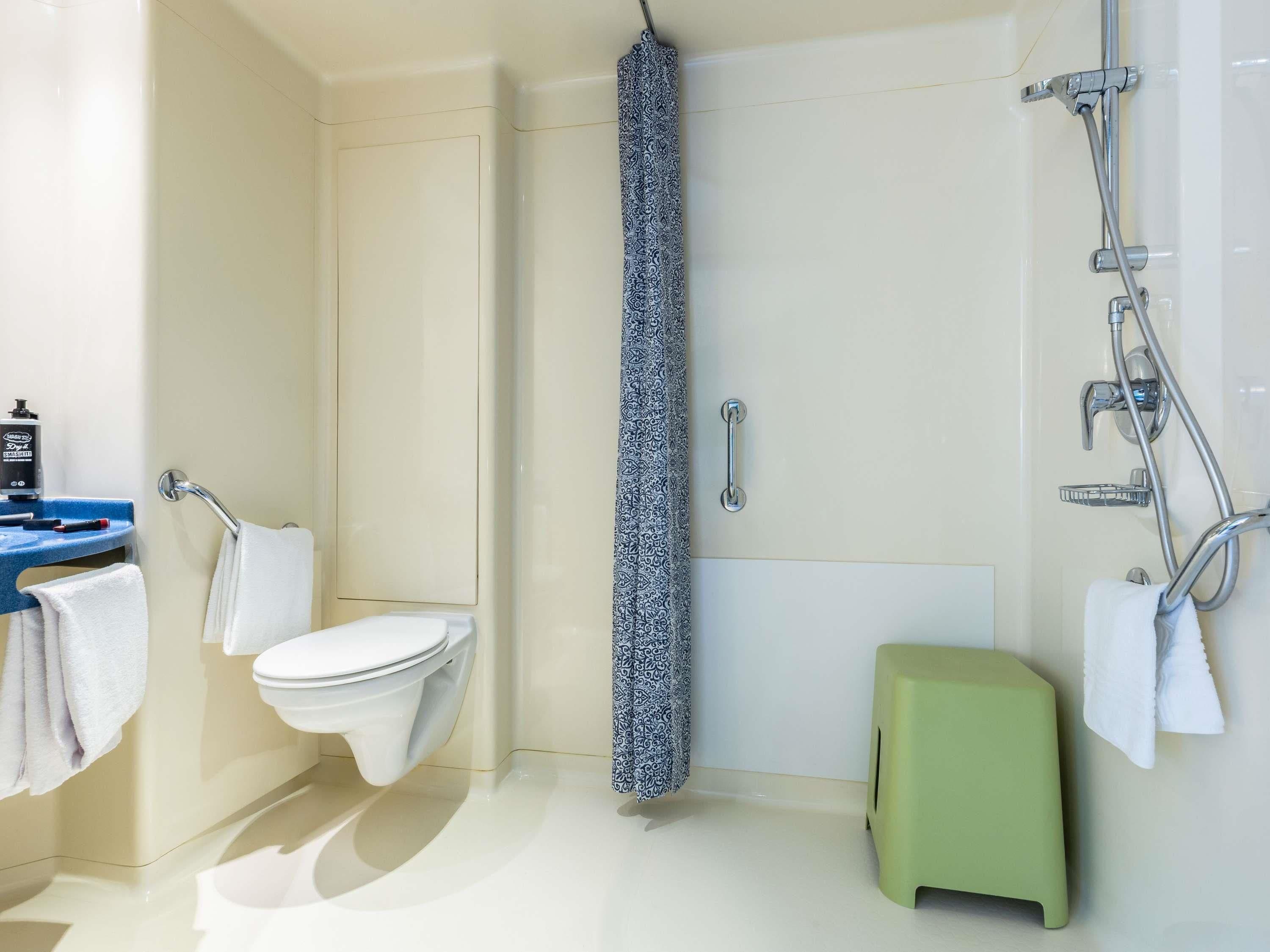 Un baño con inodoro, ducha con cortina estampada, lavamanos con toalla y un taburete verde.