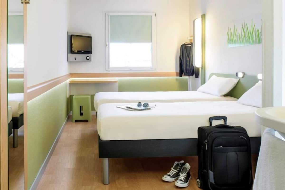A hotel room with two beds, a TV, a window, a desk, and a suitcase.
