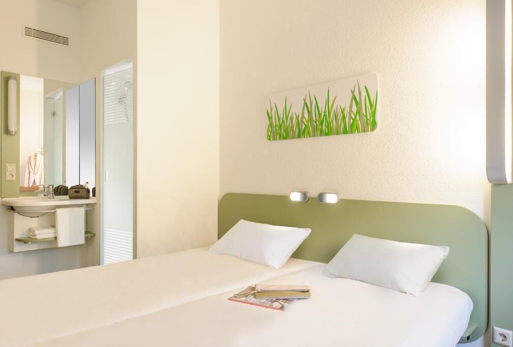 Una camera d'albergo con un letto, un comodino, uno specchio, un quadro appeso al muro raffigurante un prato e una finestra con persiane.