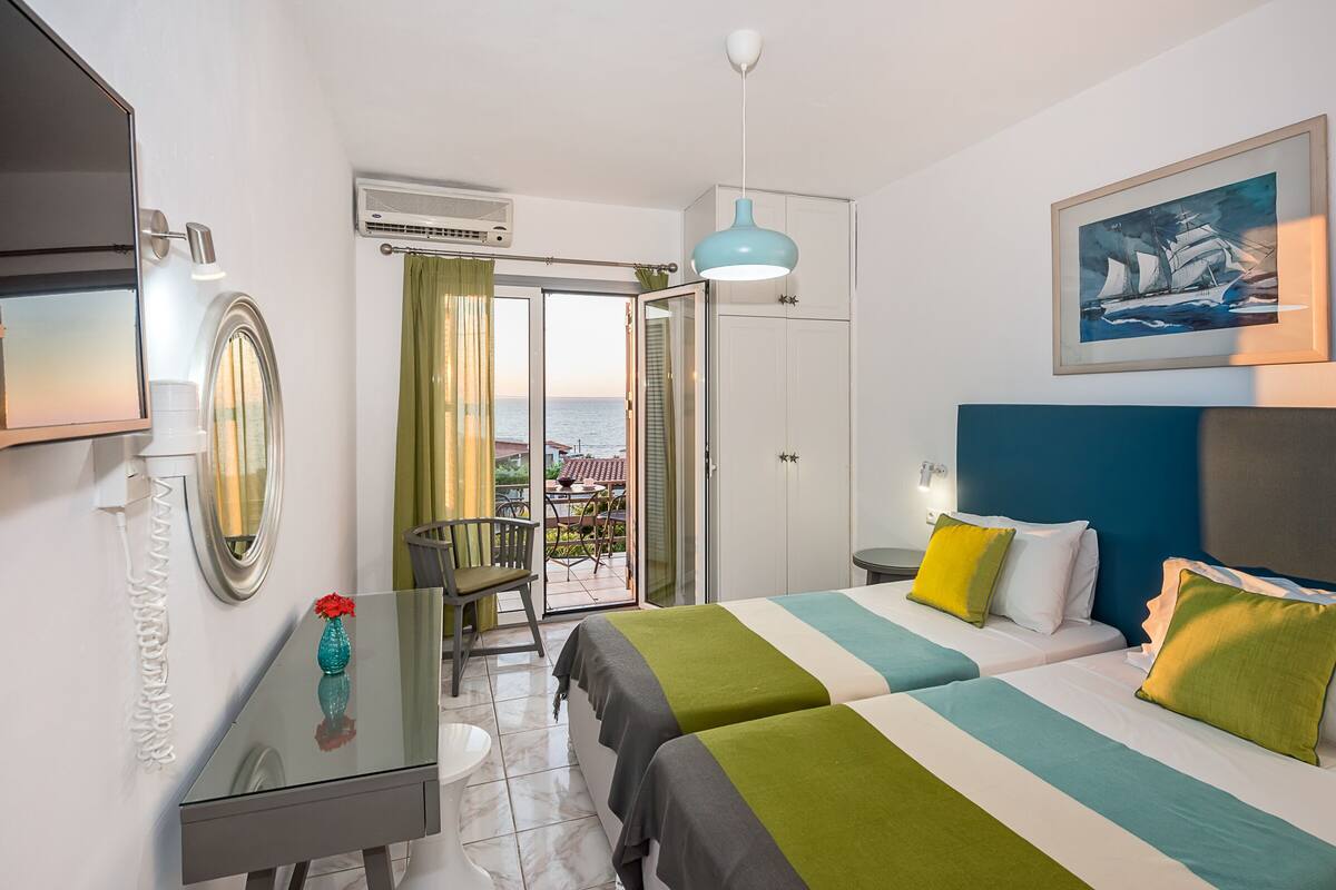A hotel room with two beds, a desk, a chair, a TV, and a view of the sea through a glass door.