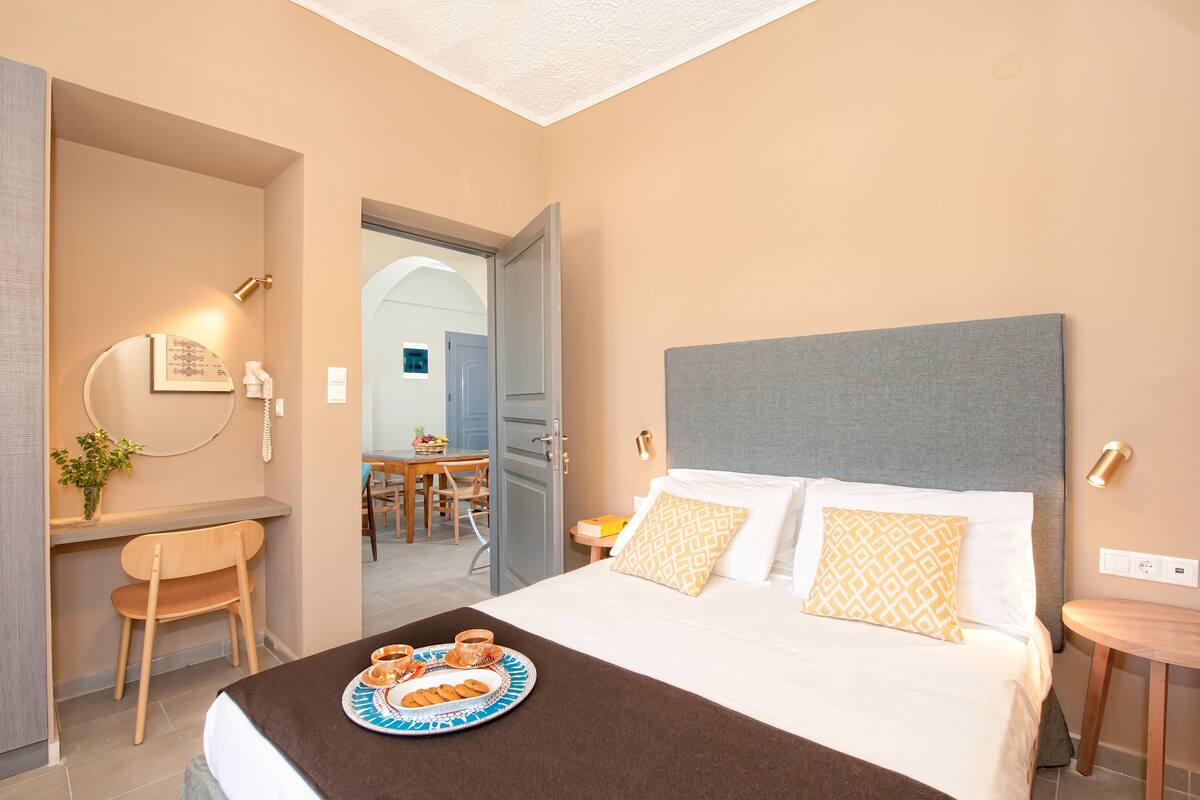 A hotel room with a bed, bedside table, and a desk with a chair. There is a dining area visible through an open door.