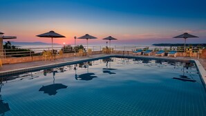 A swimming pool with sun loungers and umbrellas, surrounded by a low wall and a view of the sea at sunset. - Creta Vitalis (Chania)