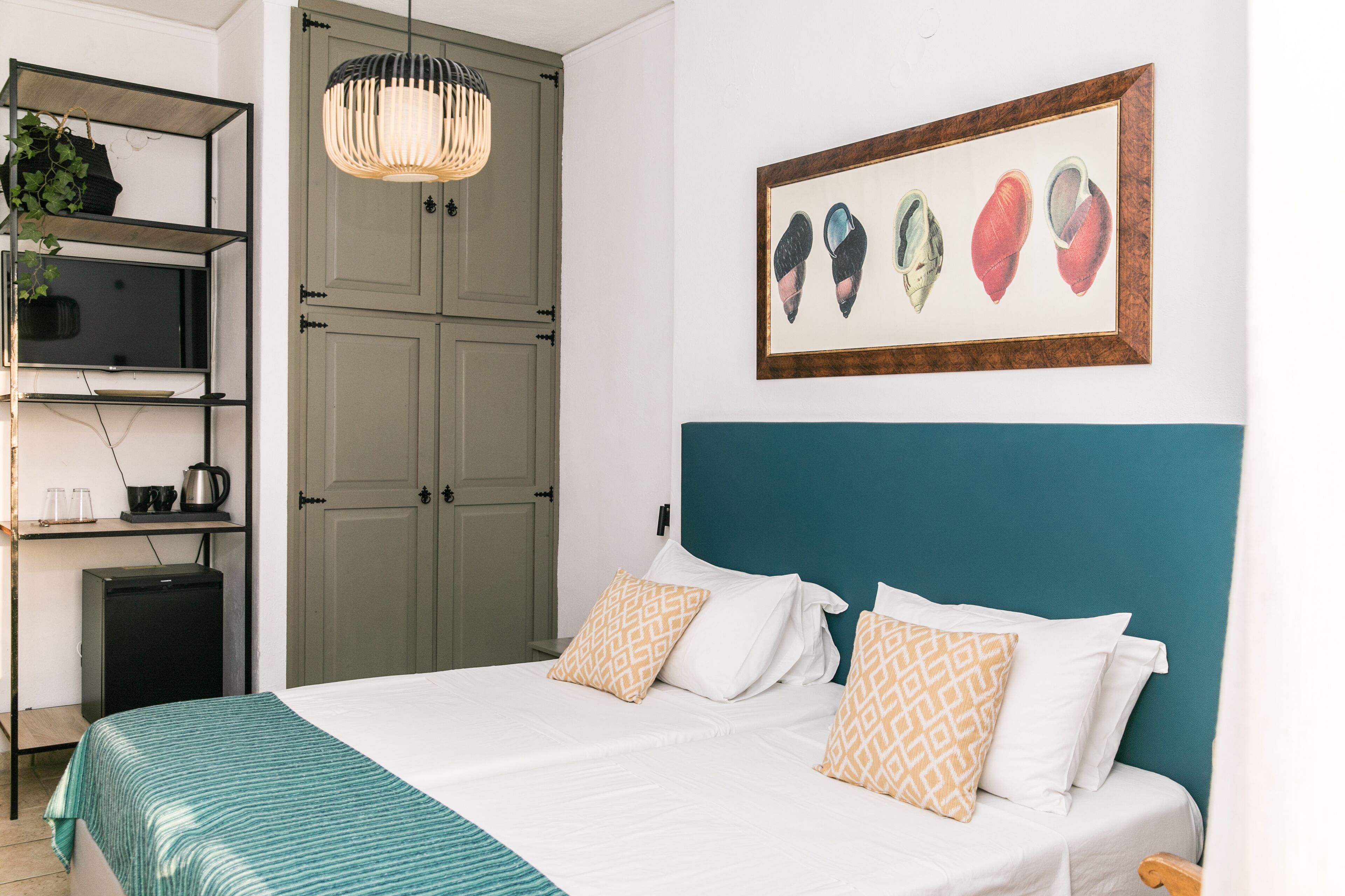 Quarto bem arrumado com uma cama, travesseiros, uma estante com uma planta, uma pequena escrivaninha com uma chaleira e um quadro na parede.