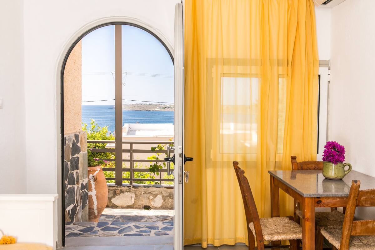 A room with a view of the sea, a dining table with chairs, a vase with flowers, and a yellow curtain.